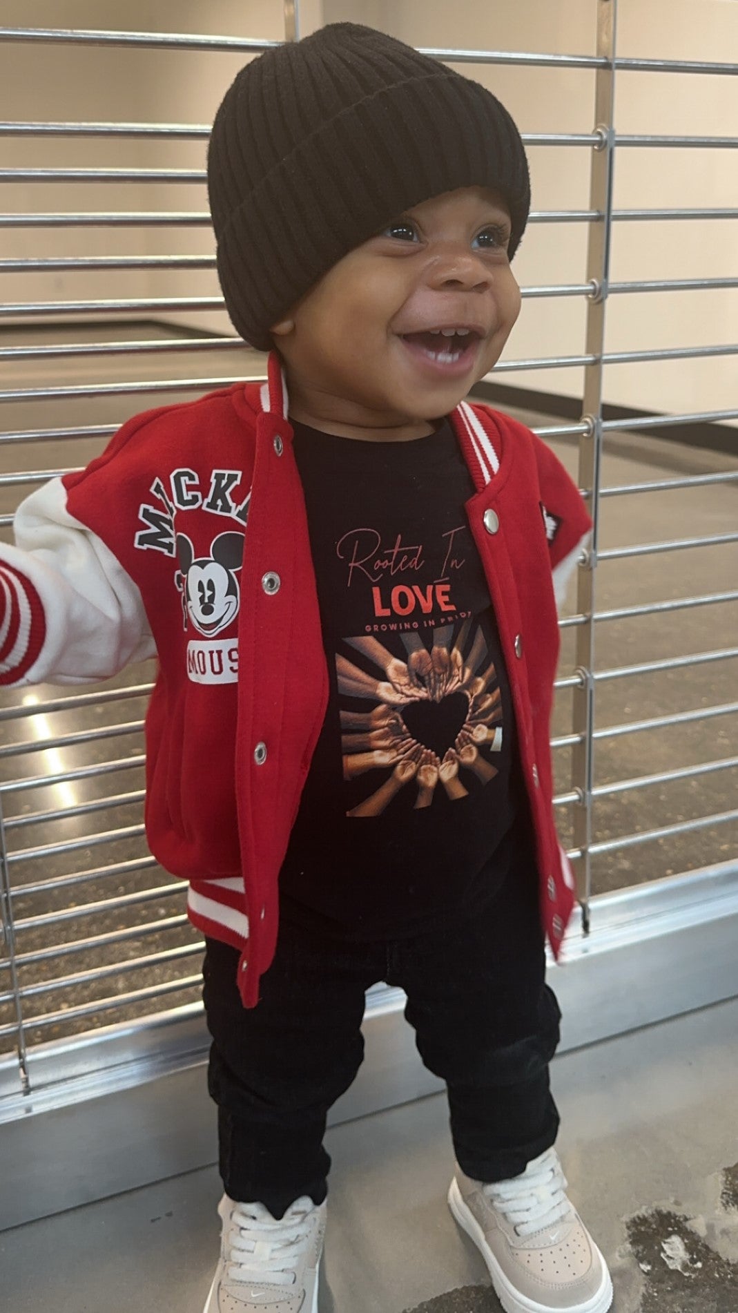 A joyful Black boy stands confidently, wearing a "Rooted in Love" t-shirt. His bright smile radiates happiness, symbolizing strength, self-love, and pride. The bold design on his shirt reflects the message that love and empowerment are everyday essentials, 