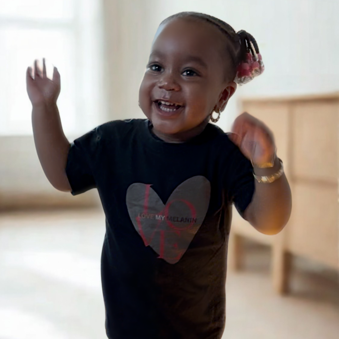 Toddler wearing Black toddler t-shirt with a large brown heart and “I Love My Melanin” text in red and brown shades, celebrating melanin pride.